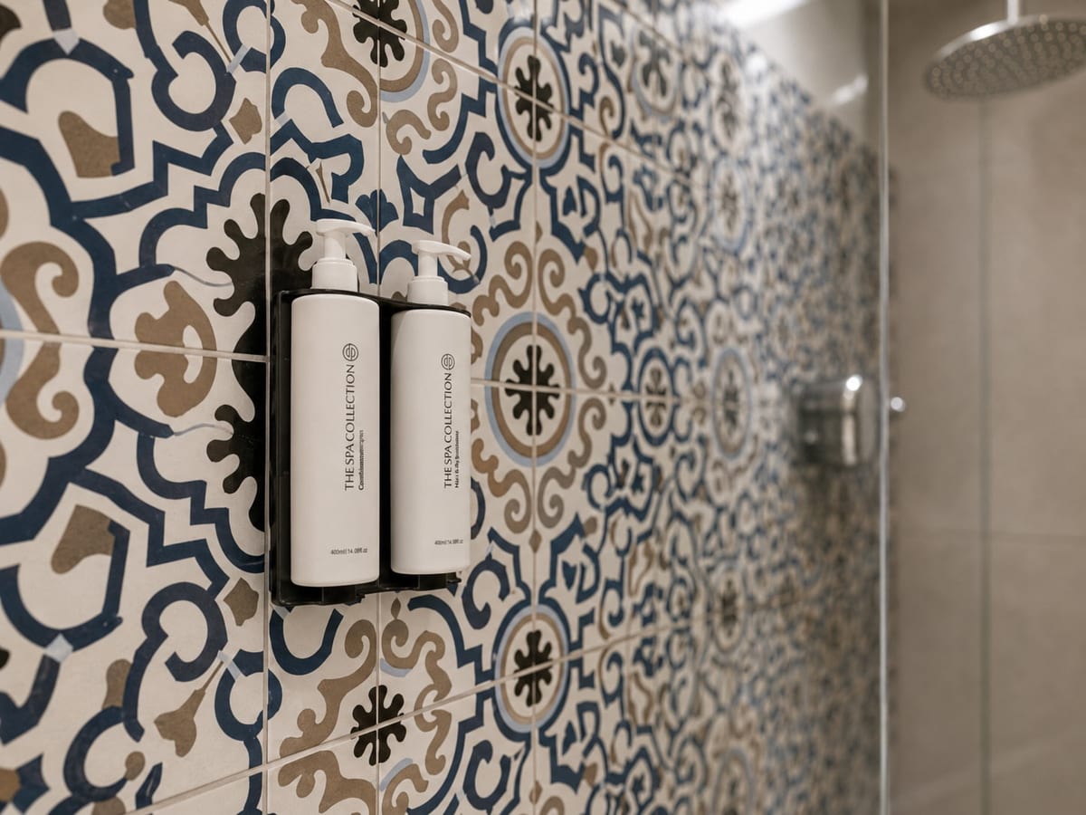 A wall of cobalt and sand majolica in the shower, with two white amenity bottles on a black bracket and the rain head behind the glass
