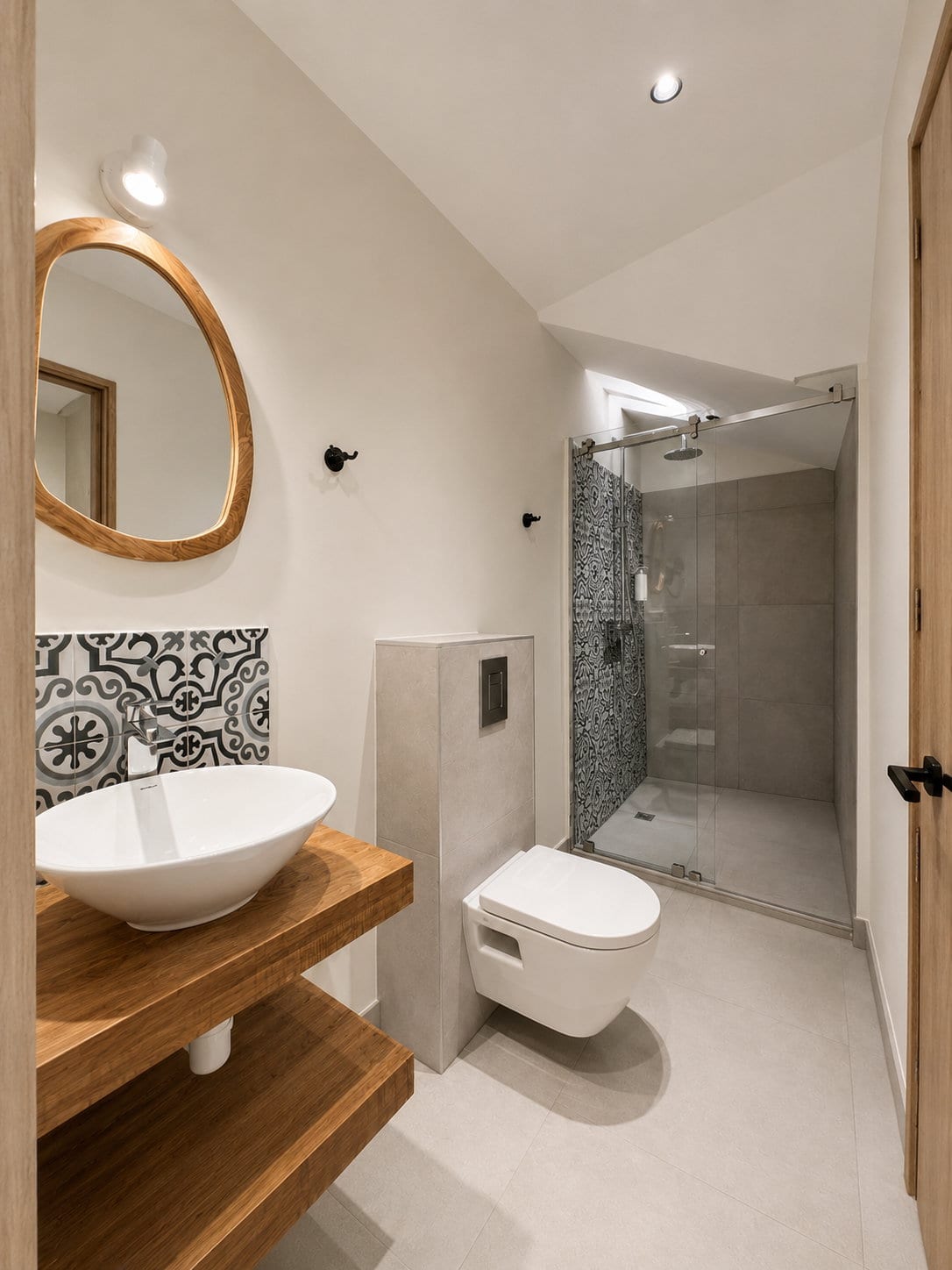 A loft bathroom with an arched mirror, a vessel basin on an oak shelf, a wall hung lavatory and a glass walk in shower lined with patterned tile