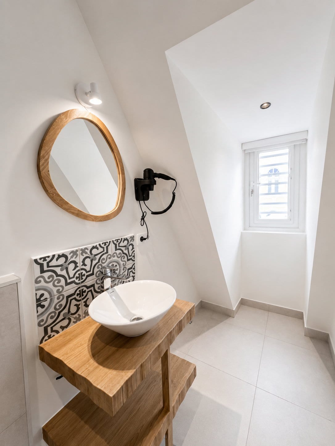 A white vessel basin on a thick oak shelf, an arched mirror above it, majolica behind and daylight from the dormer window